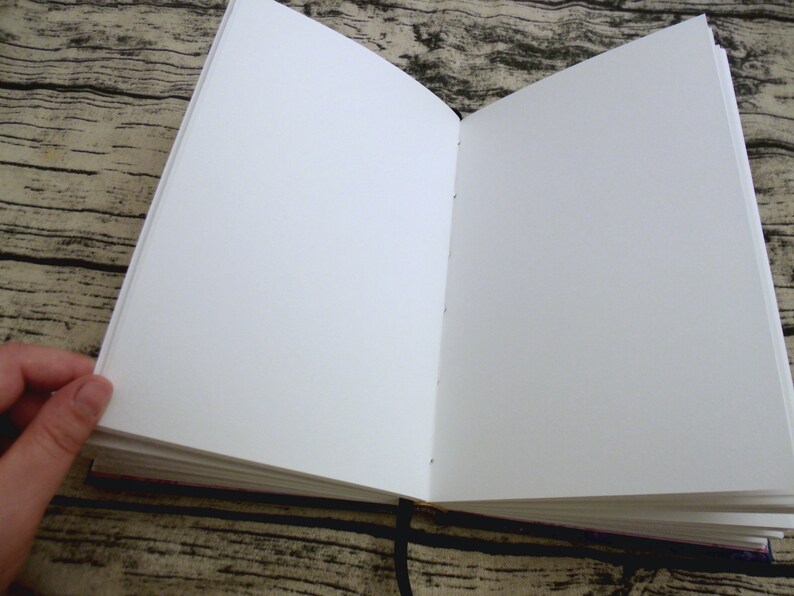May include: A blank, white, hardcover journal with a black ribbon bookmark. The journal is open to a fresh, blank page.