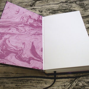 May include: An open journal with a marbled pink and white cover and blank white pages. The journal has a black ribbon bookmark.