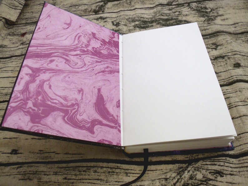 May include: An open journal with a marbled pink and white cover and blank white pages. The journal has a black ribbon bookmark.