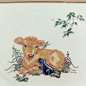 May include: A framed embroidery piece on cream fabric featuring a golden-brown calf lying down next to a small black and white skunk. The scene includes green foliage, small pink flowers, and a leafy branch above, all enclosed in a thin brown border.