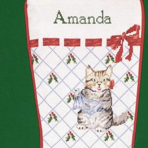 May include: A white Christmas stocking with a red trim, featuring a cross-stitched pattern of a tabby cat and a mouse. The name Amanda is stitched at the top. A red bow and holly sprigs decorate the grid design. The text Design Adapted by Jeanne Bowers is at the bottom.
