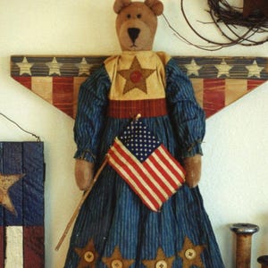 May include: A fabric teddy bear doll wearing a blue pinstriped dress with a tan bodice featuring a brown star and a red button. The skirt has four brown star appliqués with buttons and a red and tan striped hem. It holds a small American flag.
