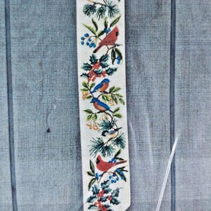 May include: A vertical fabric wall hanging featuring a cross-stitch pattern of various birds, including cardinals and bluebirds, perched among green pine branches, holly leaves, and red and blue berries. The banner has a gold rod and a white tassel.