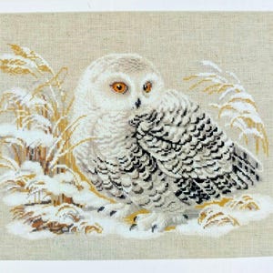 May include: A cross-stitch of a snowy owl with orange eyes, perched in a snowy landscape with wheat-colored grass. The owl has white feathers with black markings. The background is a light beige.