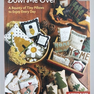 May include: A book titled Bowl Me Over: A Bounty of Tiny Pillows to Enjoy Every Day by Debbie Busby. The cover features numerous small, handcrafted fabric pillows with various embroidered designs, patterns, and text, arranged in bowls and boxes.