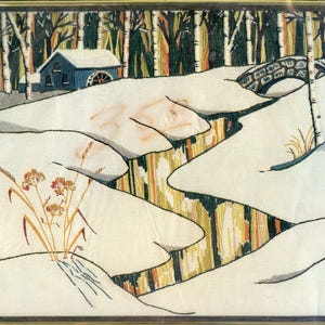 May include: A framed embroidery depicting a winter landscape with a blue house, a stone bridge, and a winding stream. The scene features white snow-covered ground, tall birch trees, and a small cluster of dried orange flowers in the foreground.