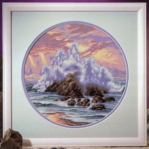 May include: A framed cross-stitch embroidery depicting a group of white horses emerging from ocean waves around a rocky outcrop. The scene features a sunset sky with shades of pink, orange, and purple, set within a circular mat and white frame.