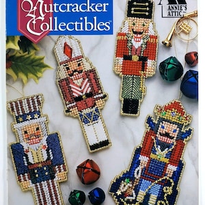 May include: A craft pattern booklet titled Nutcracker Collectibles featuring four plastic canvas nutcracker ornaments in red, white, blue, and green with gold trim, accompanied by several small metal jingle bells and a gold-toned trumpet charm.