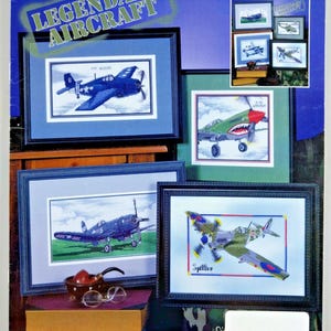 May include: A Stoney Creek Collection cross stitch pattern book titled Legendary Aircraft, featuring framed examples of embroidered airplanes including a blue F4F Wildcat, a P-40 Warhawk, and a Spitfire, with a small pipe and glasses in the foreground.