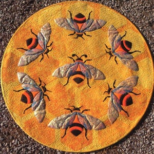 May include: A round, quilted mat with a honeycomb pattern in shades of yellow and orange. Six detailed bee appliqués are arranged around the center. The bees have orange and black bodies, and iridescent wings.