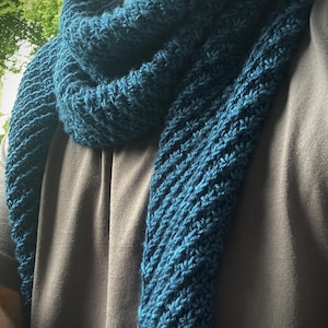 以下が含まれることがあります： テクスチャのあるパターンが施されたターコイズブルーのニットスカーフ。スカーフは折り畳まれ、人の首にかけられています。