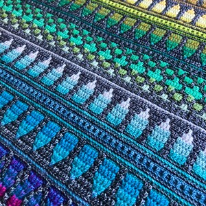 Op de afbeelding: Een close-up van een kleurrijke, gehaakte deken. De deken heeft een herhalend patroon van geometrische vormen en strepen in tinten blauw, groen, geel, oranje en paars. De textuur van het garen is zichtbaar, wat een gezellig en handgemaakt gevoel cre&euml;ert.