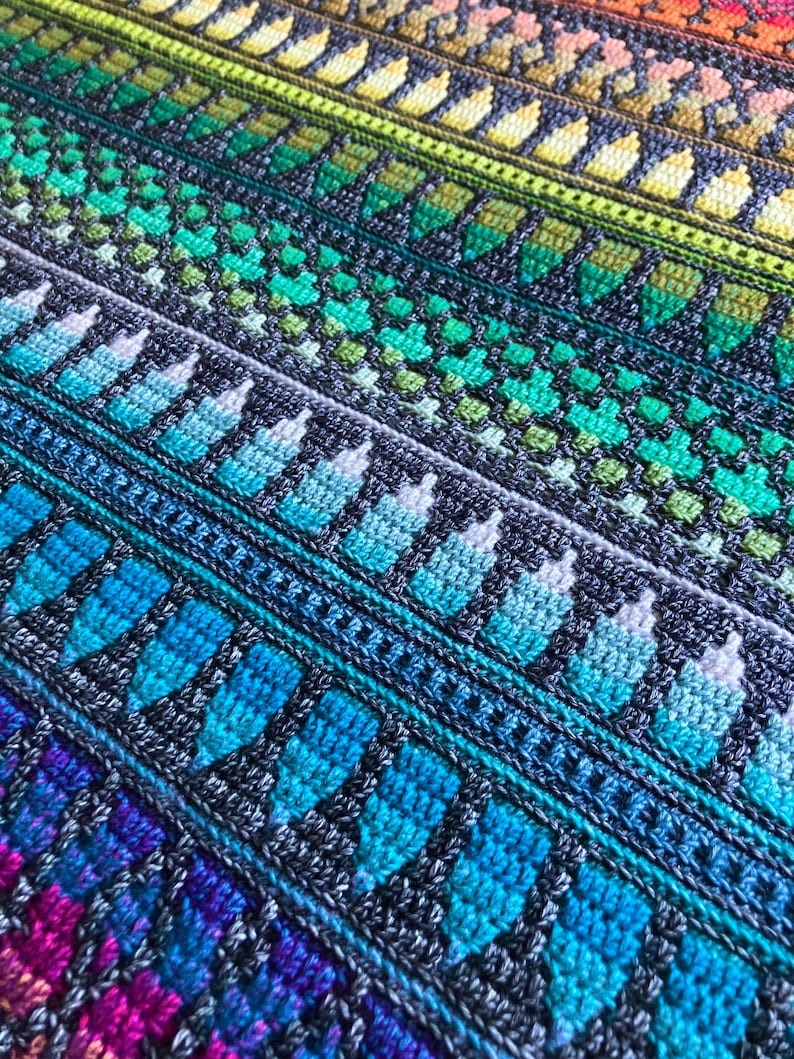 Op de afbeelding: Een close-up van een kleurrijke, gehaakte deken. De deken heeft een herhalend patroon van geometrische vormen en strepen in tinten blauw, groen, geel, oranje en paars. De textuur van het garen is zichtbaar, wat een gezellig en handgemaakt gevoel cre&euml;ert.