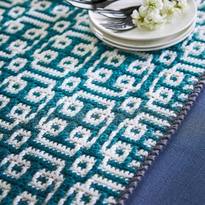 May include: A teal and white crocheted table runner with a geometric pattern. The runner is on a blue surface with white plates and cutlery on top.