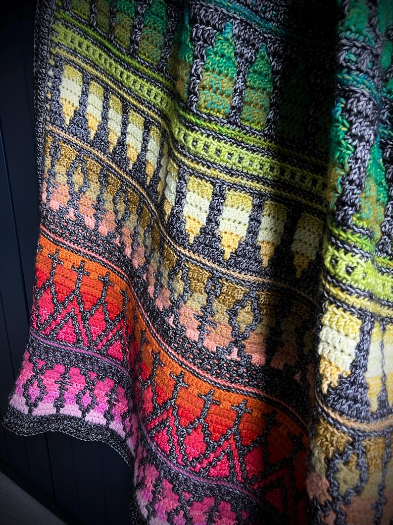 Op de afbeelding: Een kleurrijke, handgehaakte deken met een geometrisch patroon. De deken heeft een kleurengradatie, waaronder roze, rood, oranje, geel, groen en grijs. De randen zijn afgewerkt met een donkergrijze rand. De deken is opgevouwen en toont zijn textuur.