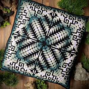 Op de afbeelding: Een vierkante gehaakte kussensloop met een complex geometrisch patroon in tinten zwart, wit en groenblauw. Het ontwerp bestaat uit een centraal stervormig motief omringd door ingewikkelde in elkaar grijpende Griekse randen van garen.
