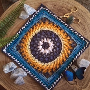 May include: A crocheted square featuring a colourful mandala design in shades of blue, yellow and white. The square is bordered with blue and white yarn.