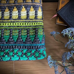 Op de afbeelding: Close-up van een kleurrijk, gehaakt textiel met een herhalend patroon van flesachtige vormen in geel, groen en blauwgroen. Het ingewikkelde ontwerp bevat geometrische en bloemelementen, waardoor een getextureerd, handgemaakt uiterlijk ontstaat. Het textiel is geplaatst op een houten oppervlak.