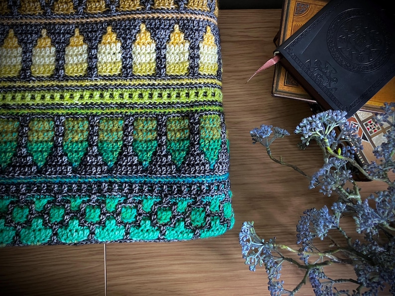 Op de afbeelding: Close-up van een kleurrijk, gehaakt textiel met een herhalend patroon van flesachtige vormen in geel, groen en blauwgroen. Het ingewikkelde ontwerp bevat geometrische en bloemelementen, waardoor een getextureerd, handgemaakt uiterlijk ontstaat. Het textiel is geplaatst op een houten oppervlak.