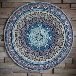 Puede incluir: Un gran mandala redondo tejido a crochet con un degradado de color azul, verde y morado. El mandala está colgado en una pared de madera.