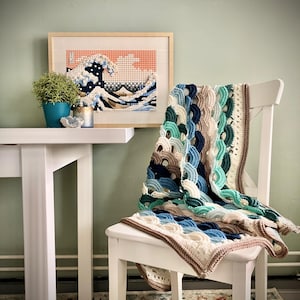 Op de afbeelding: Een handgemaakte gehaakte deken, gedrapeerd over een witte stoel. De deken heeft een golfpatroon in tinten blauw, groen en beige, met een bruine rand. Een ingelijst kunstwerk van een golf en een kleine potplant zijn ook zichtbaar.