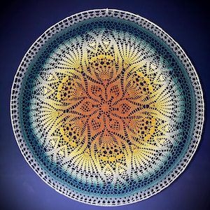 May include: A circular crochet doily featuring a vibrant gradient colour scheme that transitions from a warm orange centre to a bright yellow midsection and finishes with a deep teal blue outer edge, all enclosed within a thin white circular frame.