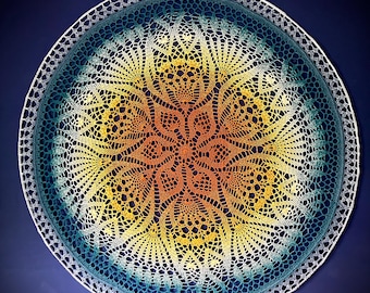 May include: A circular crochet doily featuring a vibrant gradient color scheme that transitions from a warm orange center to a bright yellow midsection and finishes with a deep teal blue outer edge, all enclosed within a thin white circular frame.