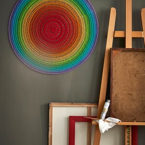 May include: A colorful rainbow crochet mandala hanging on a green wall. The mandala is made of yarn in shades of red, orange, yellow, green, blue, and purple. It is hanging on a wall next to an easel with a canvas and paint.