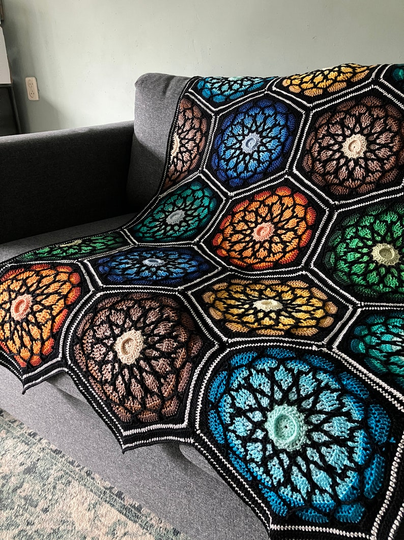 May include: A colorful crocheted blanket with a geometric pattern of hexagons. The blanket is made with a variety of shades of blue, green, orange, and brown yarn.
