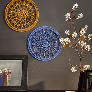 May include: Two crocheted wall hangings, one in a mustard yellow and one in a light blue, with a white flower branch in a black vase on a dark wood table.