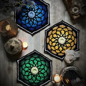 May include: Three crocheted hexagonal coasters in blue, yellow, and green with a black and white border. The coasters are on a wooden surface with candles and other decorative items.