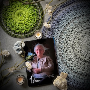 May include: A photograph of a woman with a memorial card, two crocheted doilies, candles, and decorative branches. The doilies are green and blue, with intricate openwork patterns. The memorial card has the name "Thien" and dates. The scene is arranged on a gray surface.