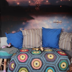 Puede incluir: Una manta de ganchillo de colores con un patrón hexagonal y una variedad de colores, incluyendo rosa, amarillo, verde y azul. La manta está drapeada sobre un sofá con almohadas y un taburete de madera.