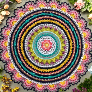 Puede incluir: Un mandala de ganchillo colorido con un diseño de múltiples capas que presenta tonos rosas, amarillos, verdes, azules y blancos. El centro del mandala tiene un diseño más pequeño e intrincado con una forma de flor.