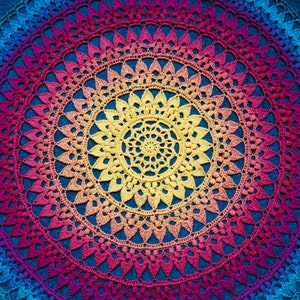 Op de afbeelding: Een kleurrijk gehaakt mandala met een regenboogkleurige gradiënt van blauw, paars, roze, oranje en geel. De mandala is gemaakt met een herhalend patroon van kleine steken en heeft een ronde vorm.