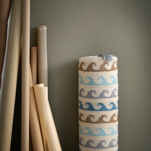 Könnte beinhalten: Ein gehäkeltes Zylinderkissen mit einem Wellenmuster in Braun-, Blau- und Grautönen. Das Kissen liegt auf einem Betonboden vor einer Wand.