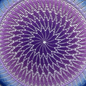 May include: A round crocheted doily with a gradient of purple, blue and white colours. The doily has a delicate, intricate design with a central point.