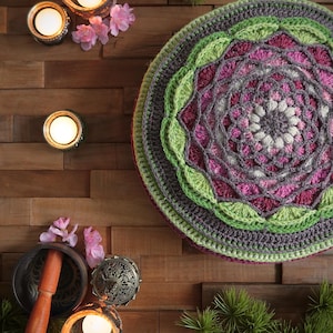 May include: A crocheted mandala meditation cushion with a pink, green, and gray color scheme. The cushion is surrounded by candles, flowers, and other meditation accessories.