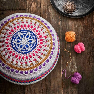 May include: A colorful crocheted floor cushion with a mandala design in shades of pink, blue, purple, and brown. The cushion is sitting on a wooden surface with balls of yarn in orange, pink, and purple.