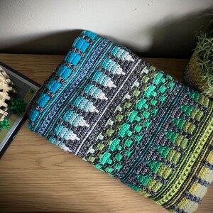 Op de afbeelding: Een kleurrijke, met de hand gehaakte deken met een geometrisch patroon. De deken heeft een combinatie van blauwe, turquoise, groene en grijze tinten. Het ontwerp bevat vierkanten, bladvormige vormen en gestructureerde strepen. De deken is op een houten oppervlak gevouwen.