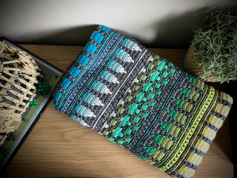Op de afbeelding: Een kleurrijke, met de hand gehaakte deken met een geometrisch patroon. De deken heeft een combinatie van blauwe, turquoise, groene en grijze tinten. Het ontwerp bevat vierkanten, bladvormige vormen en gestructureerde strepen. De deken is op een houten oppervlak gevouwen.