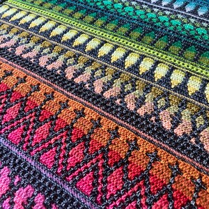 Op de afbeelding: Een close-up van een kleurrijke, gebreide deken met een kleurengradient. De deken heeft rijen ingewikkelde patronen in tinten rood, oranje, geel, groen, blauw en paars. Het ontwerp bevat ruit- en bladachtige vormen.