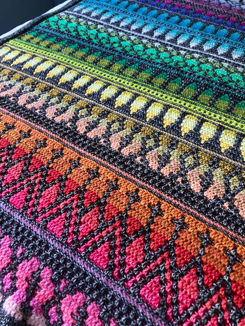 Op de afbeelding: Een close-up van een kleurrijke, gebreide deken met een kleurengradient. De deken heeft rijen ingewikkelde patronen in tinten rood, oranje, geel, groen, blauw en paars. Het ontwerp bevat ruit- en bladachtige vormen.