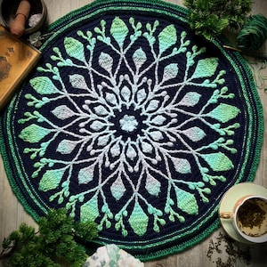 Op de afbeelding: Een ronde, handgemaakte gehaakte vloerkleed met een botanisch ontwerp. Het vloerkleed heeft een donkerblauwe kern met een wit en groen bladpatroon. De rand is groen. Andere objecten omringen het vloerkleed, waaronder een theekopje en een kristal.