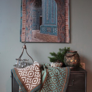 May include: A brown and teal crocheted blanket draped over a dark gray metal dresser with two drawers. A silver metal lantern with glass jars sits on top of the dresser. A decorative lantern with a gold finish sits on the dresser. A framed print of a blue and white tiled archway hangs on the wall behind the dresser.