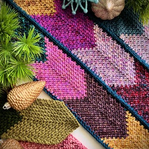 May include: A colorful crocheted scarf with a geometric pattern in shades of pink, purple, teal, and gold. The scarf is displayed with decorative ornaments.