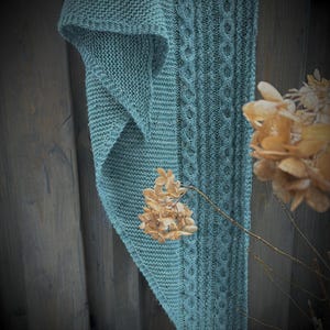 May include: A teal knitted scarf with a cable knit design and a textured stitch pattern. The scarf is draped over a wooden fence, with dried flowers in the foreground. The scarf's color is a muted, dusty teal.
