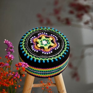 May include: A black crocheted stool with colorful stripes and a floral design. The stool has wooden legs and is sitting on a gray surface.
