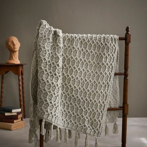 Może przedstawiać: Ręcznie robiony koc z dzianiny z teksturalnym wzorem i frędzlami. Koc jest położony na drewnianym stojaku.