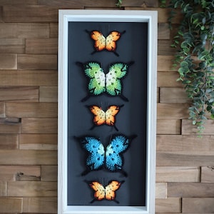 Op de afbeelding: Een wit frame met een zwarte achtergrond toont vijf gehaakte vlinders in een verticale rij. De vlinders zijn in tinten oranje, groen, blauw en geel.